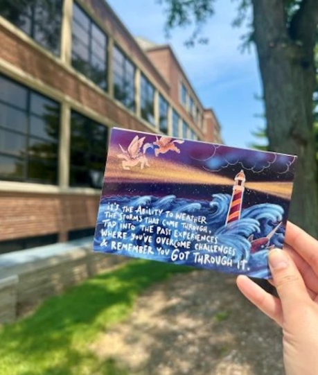A person holding up a colorful postcard outside. The postcard shows a scene of a lighthouse amidst waves. The text reads 